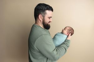 Bearded man in green sweatshirt tenderly holds sleeping newborn baby wrapped in blue blanket against beige background.
