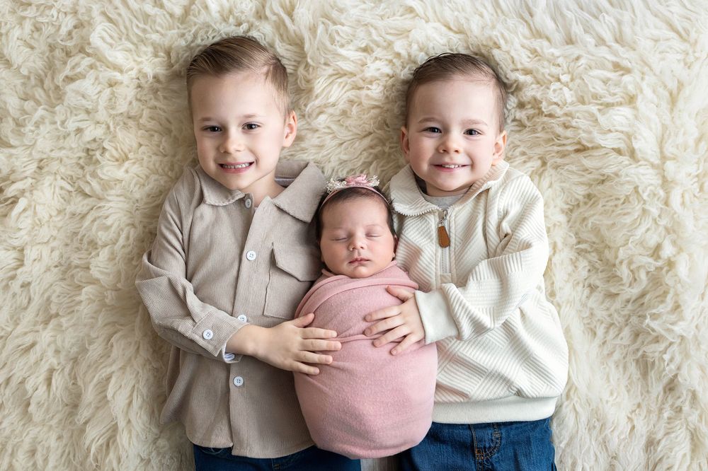 Two smiling young boys lying on a fluffy rug while gently holding their newborn baby sister wrapped in pink.