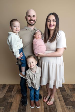 Happy family of five posing for a portrait with newborn baby, two young boys, and parents on wood floor.