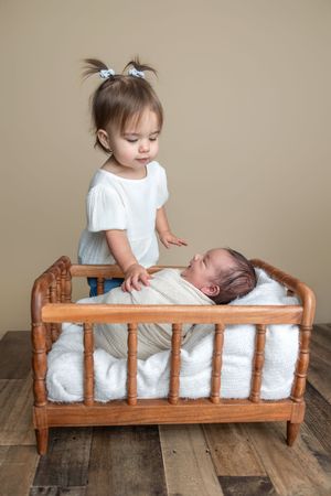 Toddler girl with pigtails standing beside a wooden crib with a sleeping newborn baby wrapped in white.
