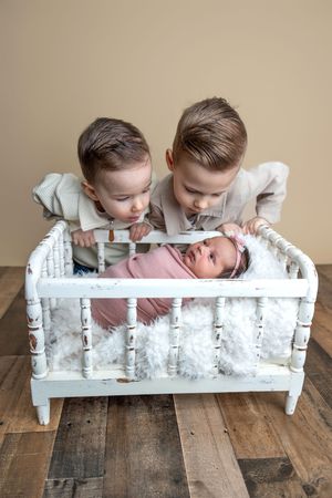 Two young boys lean over a white vintage crib to look at their newborn baby sister wrapped in pink.
