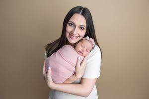 A smiling mother in a white top holds her sleeping newborn baby wrapped in a pink swaddle against a beige background.