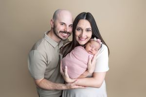 A smiling couple poses with their newborn baby wrapped in pink against a warm beige studio background.