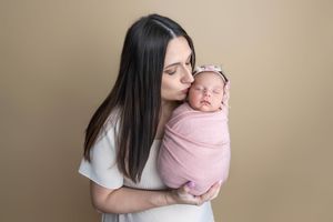 A mother kisses her sleeping newborn baby wrapped in a pink swaddle blanket against a beige background.