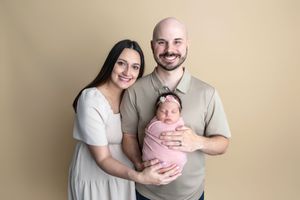 A smiling couple poses for a newborn portrait, holding their pink-swaddled baby girl against a neutral beige background.