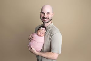 Smiling bald man holding a sleeping newborn baby swaddled in pink wrap with floral headband against beige background.