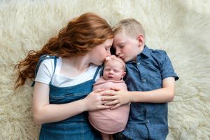 A red-haired girl and young boy lying on a fluffy rug, lovingly kissing a swaddled newborn baby.