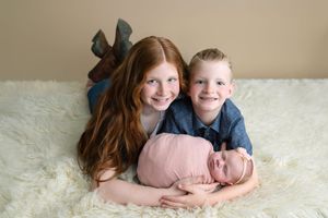 Two smiling children with red and blonde hair pose with a sleeping newborn wrapped in pink on a white fur rug.