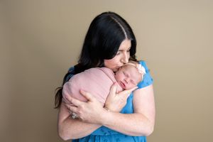 A mother with dark hair in a blue dress tenderly kisses her sleeping newborn wrapped in a pink swaddle.