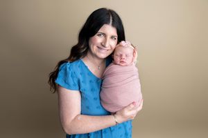 A smiling woman in a blue dress holds a sleeping newborn swaddled in pink against a neutral background.
