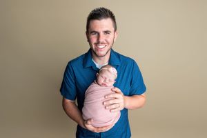 Smiling man in blue shirt holding a swaddled newborn baby wrapped in pink against a neutral background.