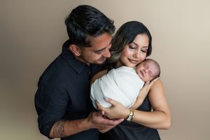 A couple in a studio portrait tenderly holds their swaddled newborn baby against a warm beige background.