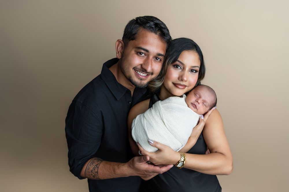 A couple in black outfits smiles warmly while cradling their swaddled newborn baby against a neutral beige backdrop.