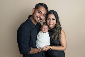 A smiling couple in black outfits holds their swaddled newborn baby during a professional studio portrait session.