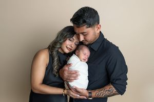 Parents in black outfits tenderly hold their swaddled newborn baby during a professional studio portrait session.