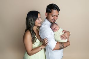 Young couple in neutral studio holding swaddled newborn baby, woman in yellow dress, man in white shirt with tattoo.