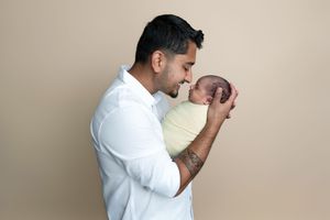A father tenderly holds his newborn baby close, sharing a gentle moment against a neutral beige background.