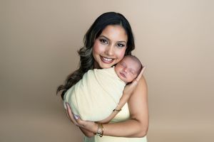 Smiling mother in yellow dress holds swaddled newborn baby during a professional studio portrait.