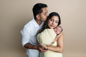 A couple tenderly holds their swaddled newborn baby, with the father kissing the mother's forehead in a studio portrait.