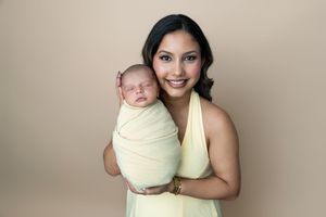 Smiling woman in yellow dress holding a swaddled newborn baby against a beige background.