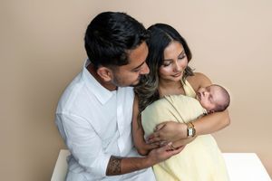 Happy couple in beige studio admiring their swaddled newborn, woman in yellow dress, man in white shirt with arm tattoo.