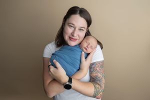 A smiling mother with a tattoo sleeve holds her sleeping newborn wrapped in a blue swaddle against a tan background.