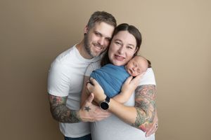 A smiling couple with tattoos poses together holding their sleeping newborn baby dressed in blue against a tan background.