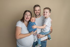Family of four posing for studio portrait with newborn baby and toddler son against beige background.