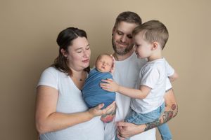 Family of four with tattooed parents in white shirts, mom holding swaddled newborn, dad holding toddler son, on beige background.
