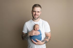 Tattooed father in white t-shirt smiling while holding sleeping newborn baby wrapped in blue swaddle against beige background.