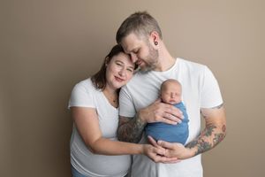 A couple in white t-shirts tenderly holds their newborn baby wrapped in blue against a neutral background.