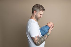 Tattooed father in white t-shirt tenderly holds sleeping newborn baby wrapped in blue swaddle against neutral background.