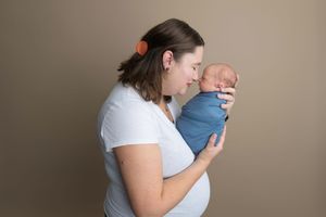 A mother in a white shirt tenderly holds her swaddled newborn baby in a blue wrap against a neutral background.