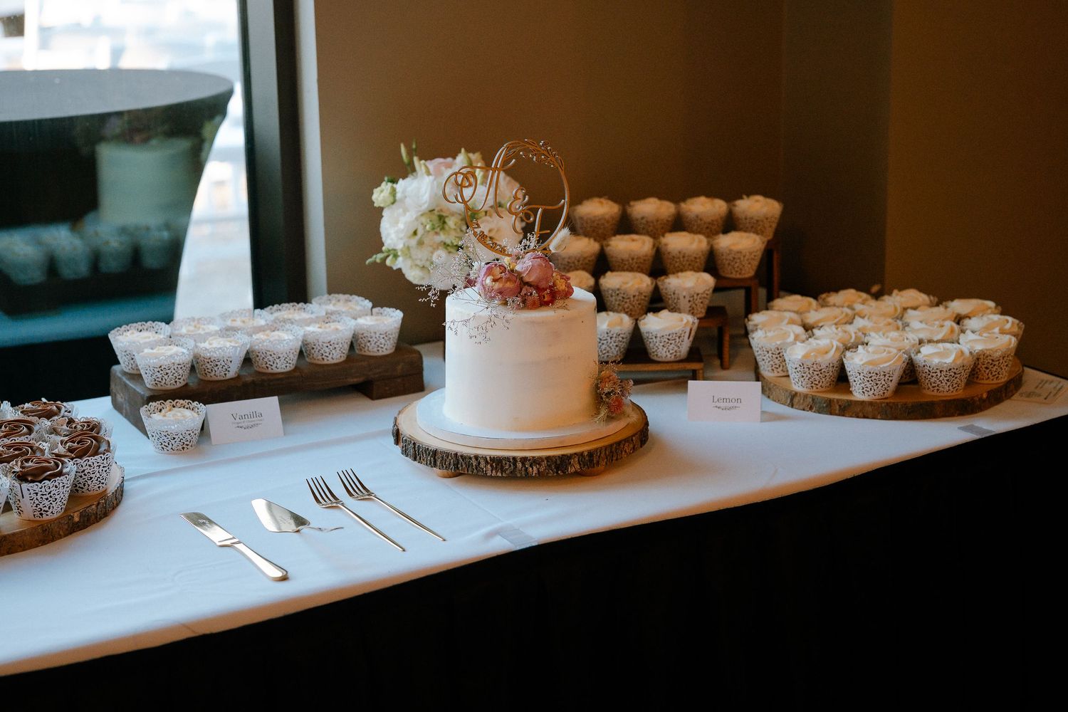 Elegant wedding dessert table with white cake on wood slice, cupcakes, and floral topper by a window.