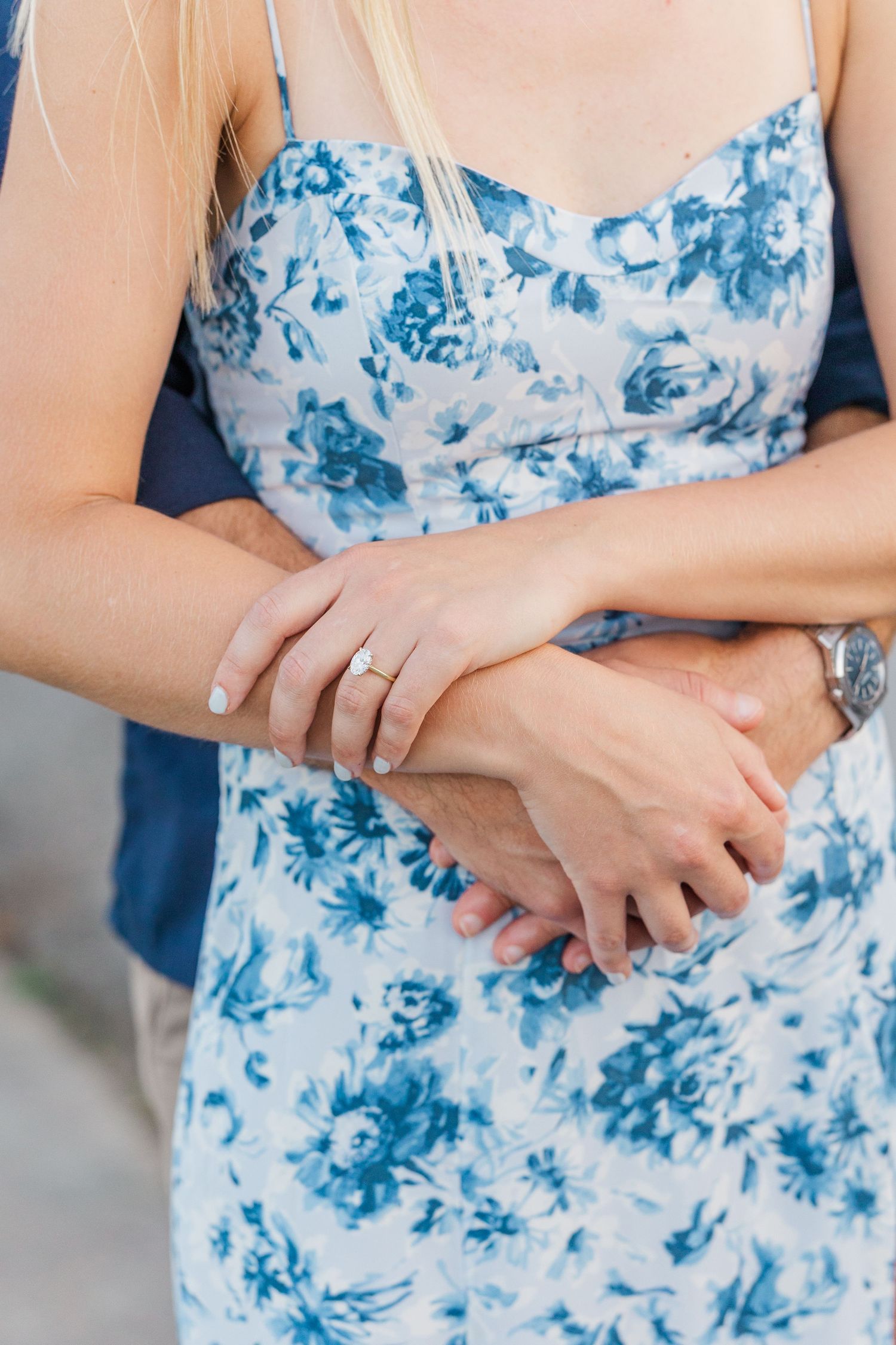 Close-up of couple holding hands, woman wearing diamond engagement ring and blue floral dress.