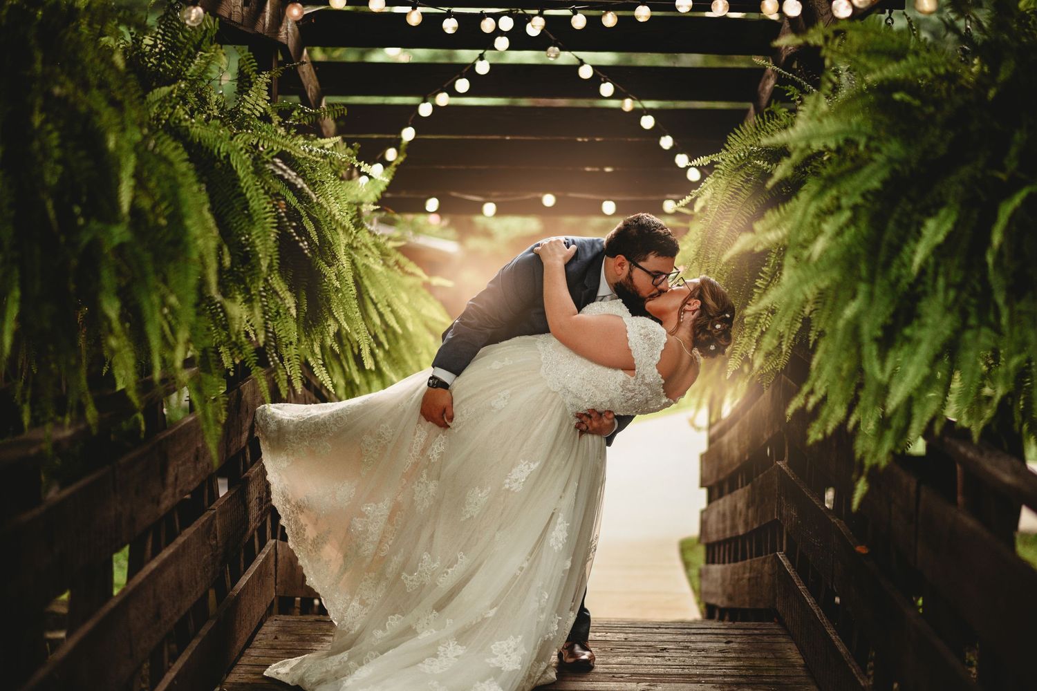 Groom dips bride in lace gown under string lights in fern-lined wooden tunnel at romantic wedding.