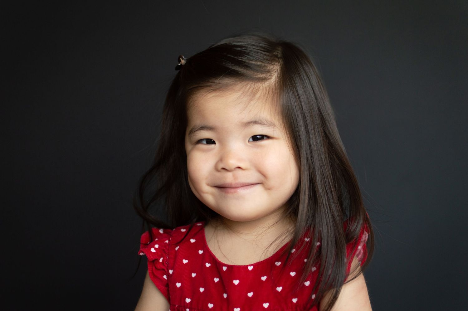 A smiling young Asian girl with long dark hair wearing a red heart-print dress poses against a dark background.