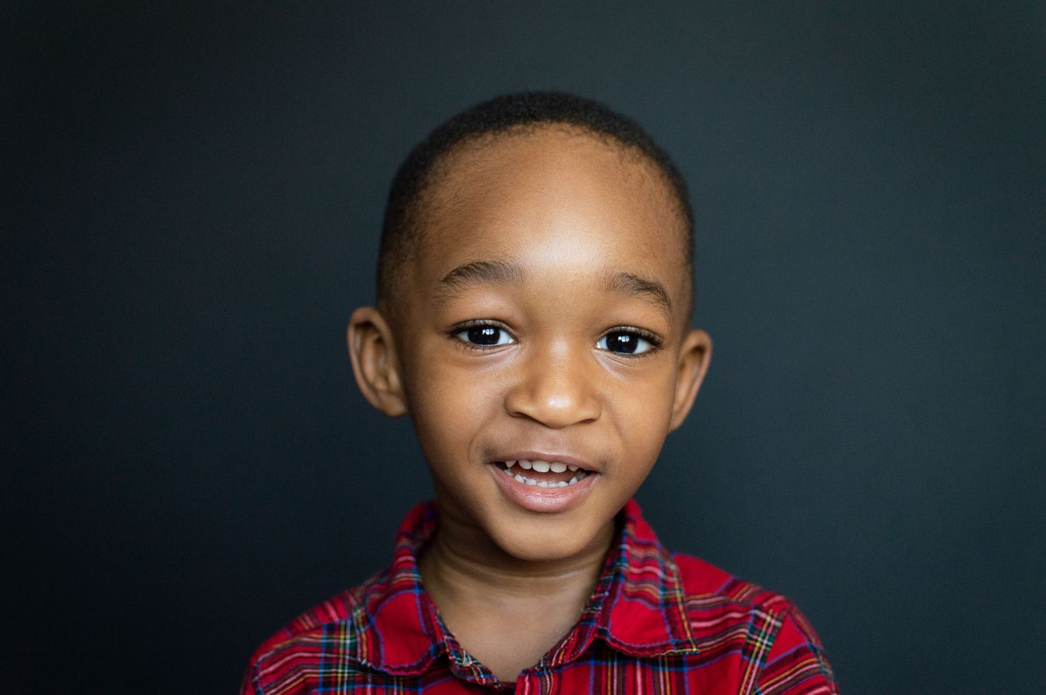 A cheerful young Black boy in a red plaid shirt smiles brightly against a dark background.