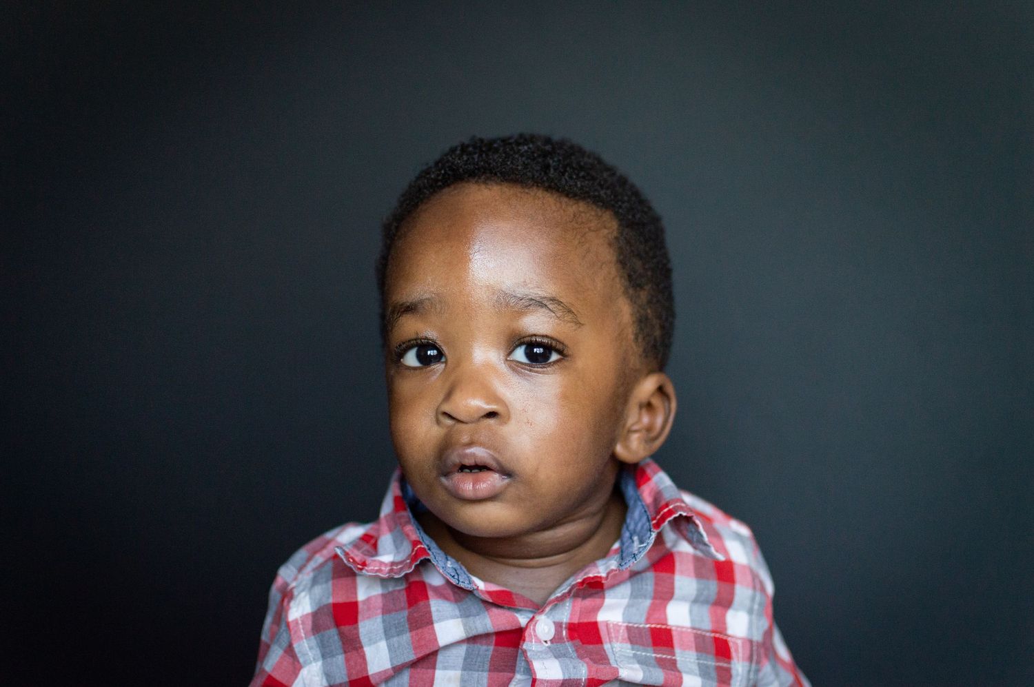 A young Black toddler boy in a plaid shirt looks curiously at the camera against a dark background.
