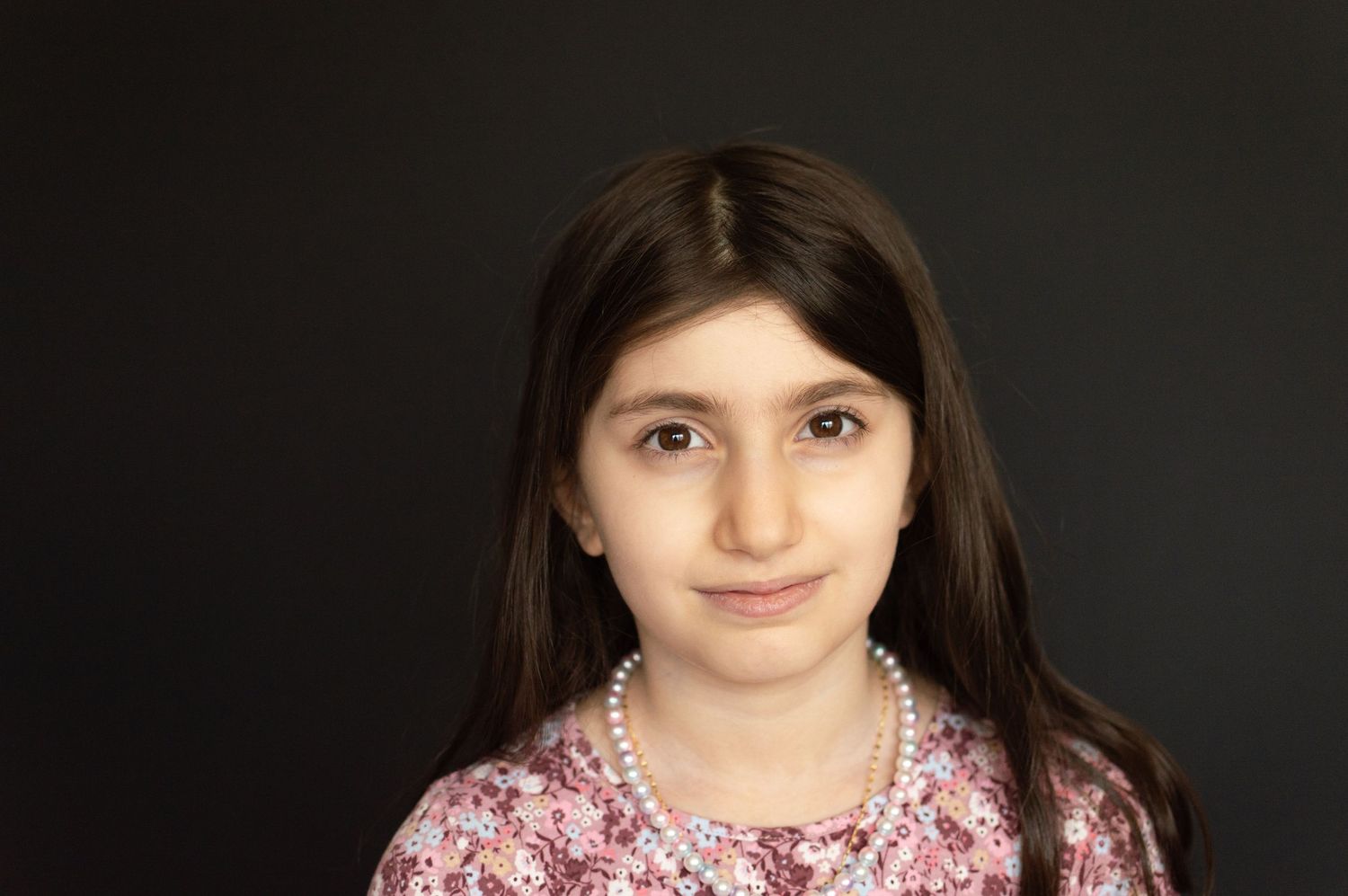 A young girl with long dark hair wearing a pearl necklace and floral top poses against a dark background.