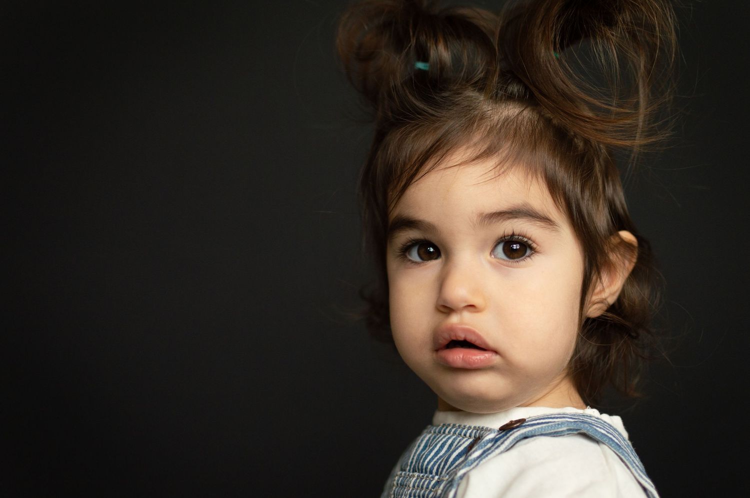 A cute toddler girl with hair in two buns wearing denim overalls glances over her shoulder on dark background.