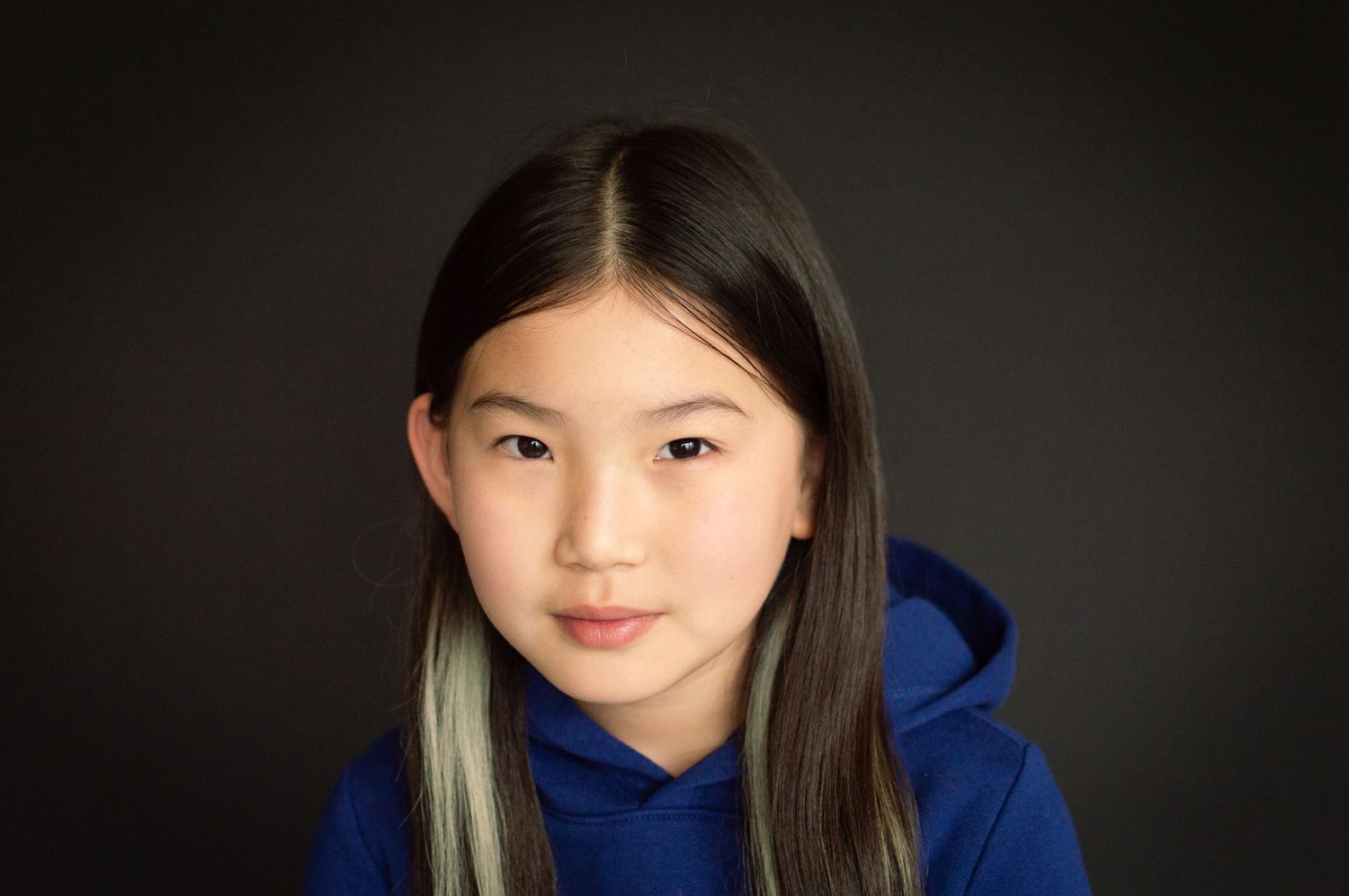 A preteen Asian girl with long dark hair and a blue streak wears a blue hoodie against a dark background.