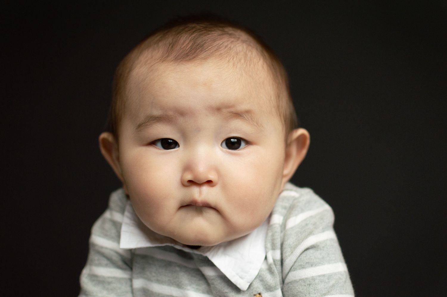 An adorable Asian baby in a gray striped outfit gazes seriously at the camera against a dark background.