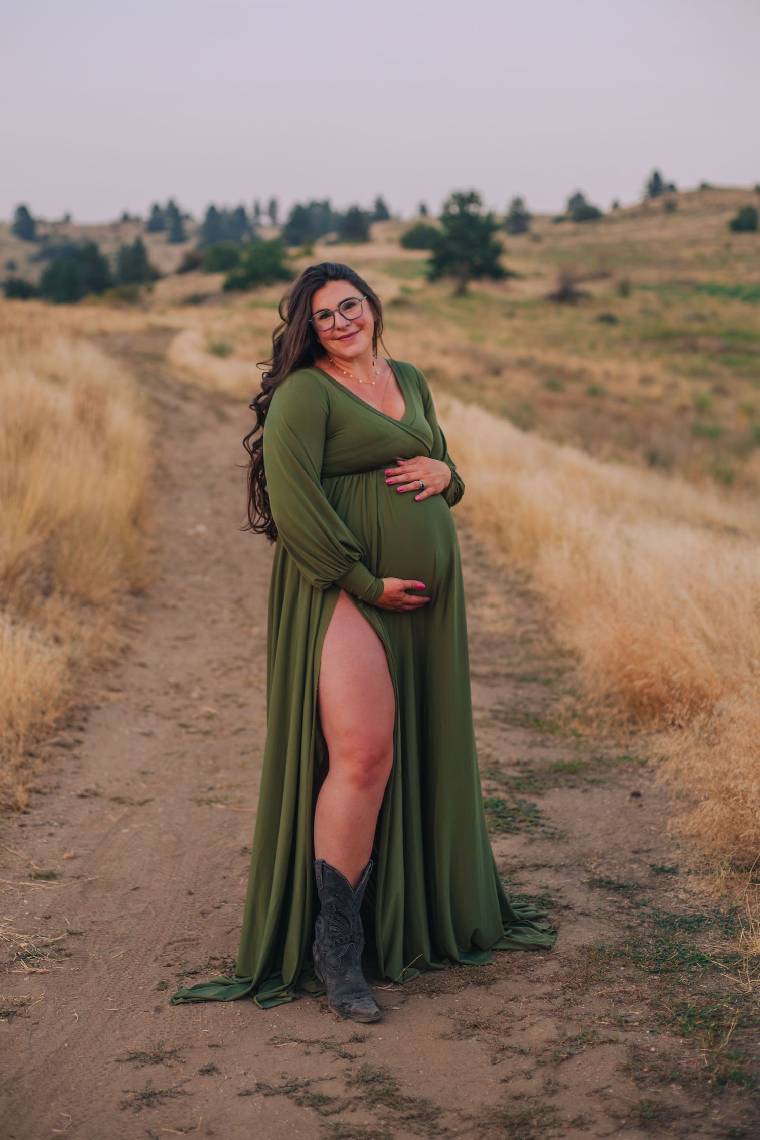Pregnant woman in green maternity gown with slit and cowboy boots poses on a dirt path in a golden field.