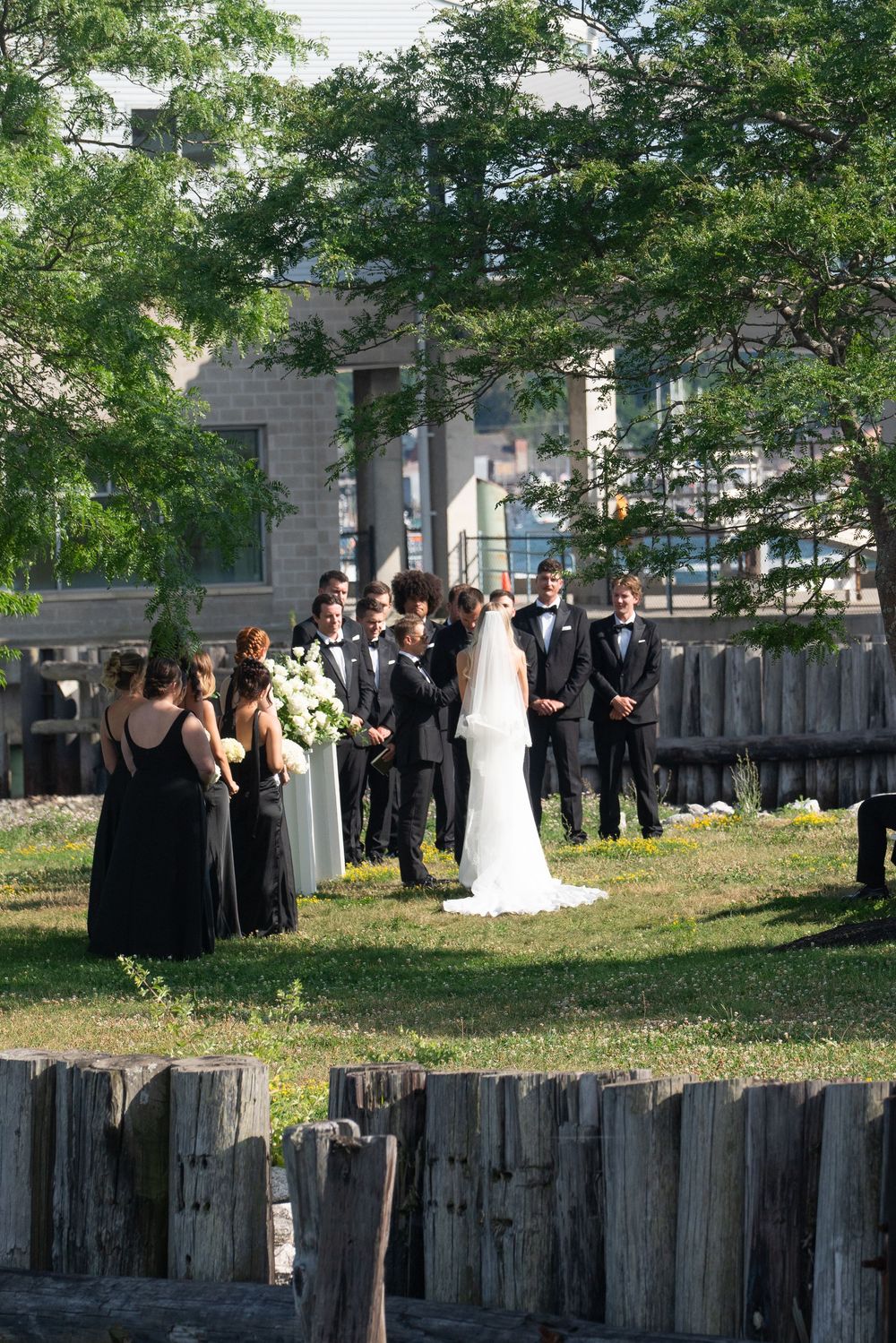 Wedding ceremony in progress outdoors with bridal party in black attire beside rustic wooden pilings.