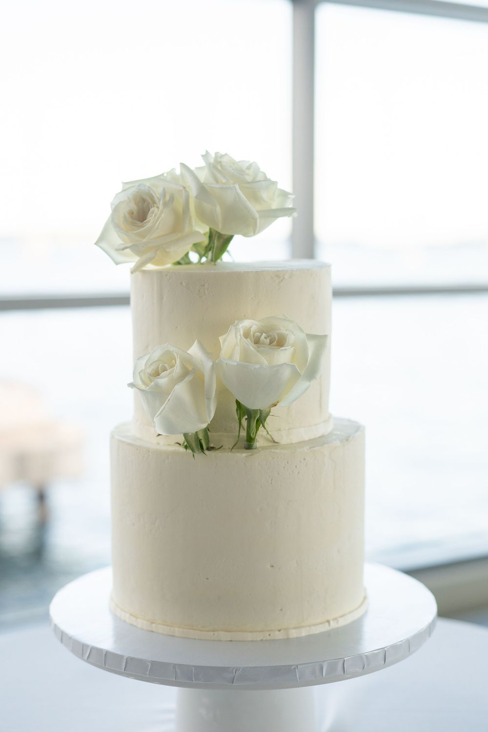 Two-tier white wedding cake decorated with fresh white roses on a white cake stand.