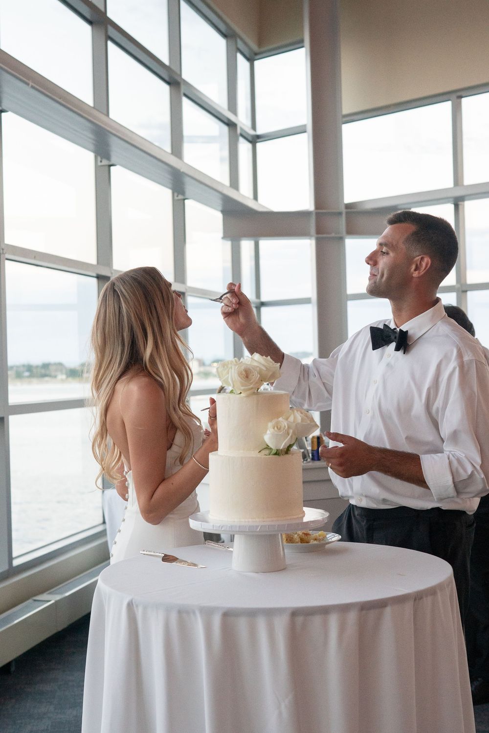 Groom feeds bride a bite of elegant two-tier white wedding cake with floral decorations.