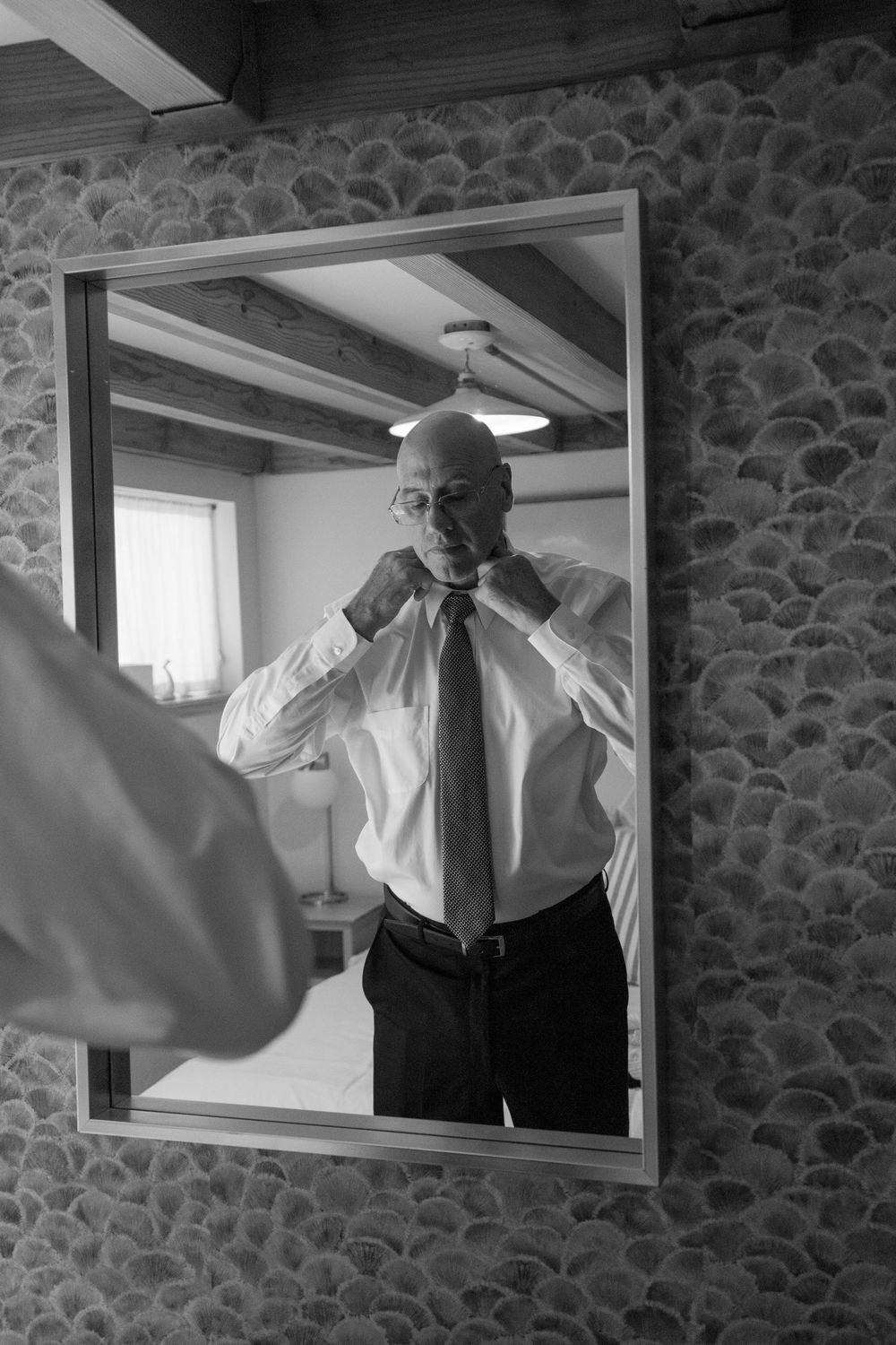 Older man adjusting his tie while looking at his reflection in a mirror before a wedding.
