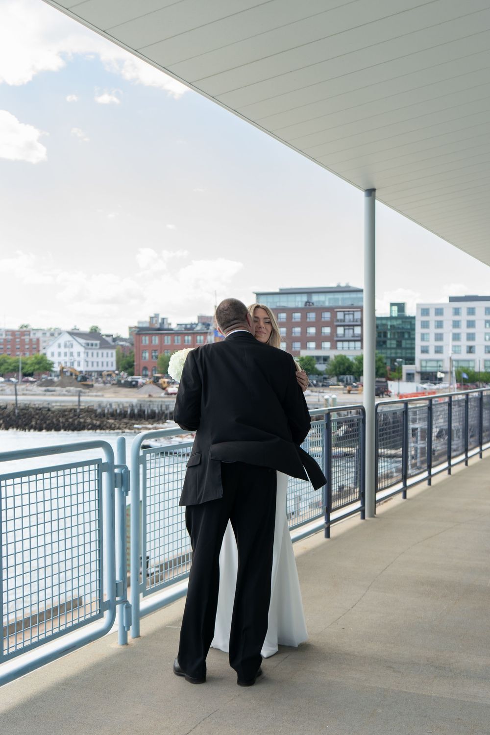 Bride and groom share a romantic moment on a waterfront promenade with city buildings in the background.