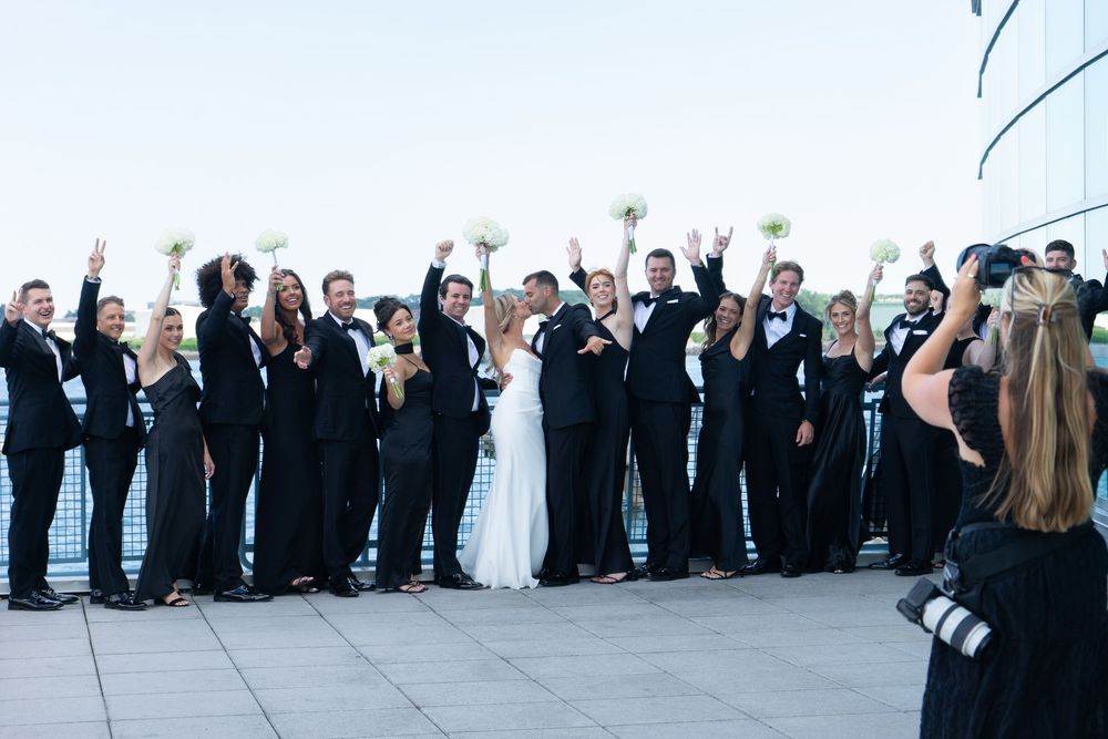 Large wedding party in black attire celebrating with raised arms at waterfront venue.